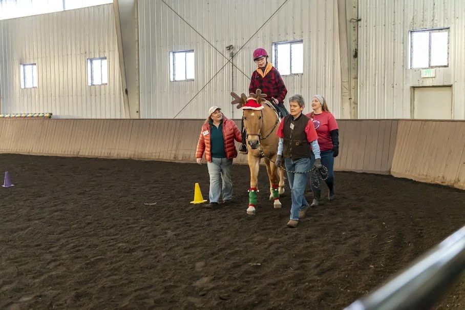 A person with Valley Girl style, wearing a red helmet and a red and black plaid shirt, riding a horse decorated with Christmas accessories inside an indoor riding arena. Three smiling women walk beside the horse, one wearing a red jacket, another in a black vest, and the third in a pink sweatshirt. Cones and small obstacles are set up on the dark dirt ground.