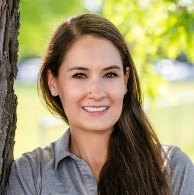 A woman with long brown hair and a gray shirt standing outdoors next to a tree, smiling with a blurred green and sunny background.