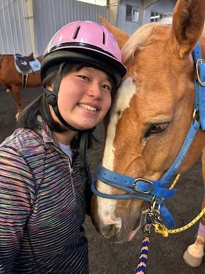 Young girl in a pink riding helmet smiling next to a light brown horse wearing a blue halter.