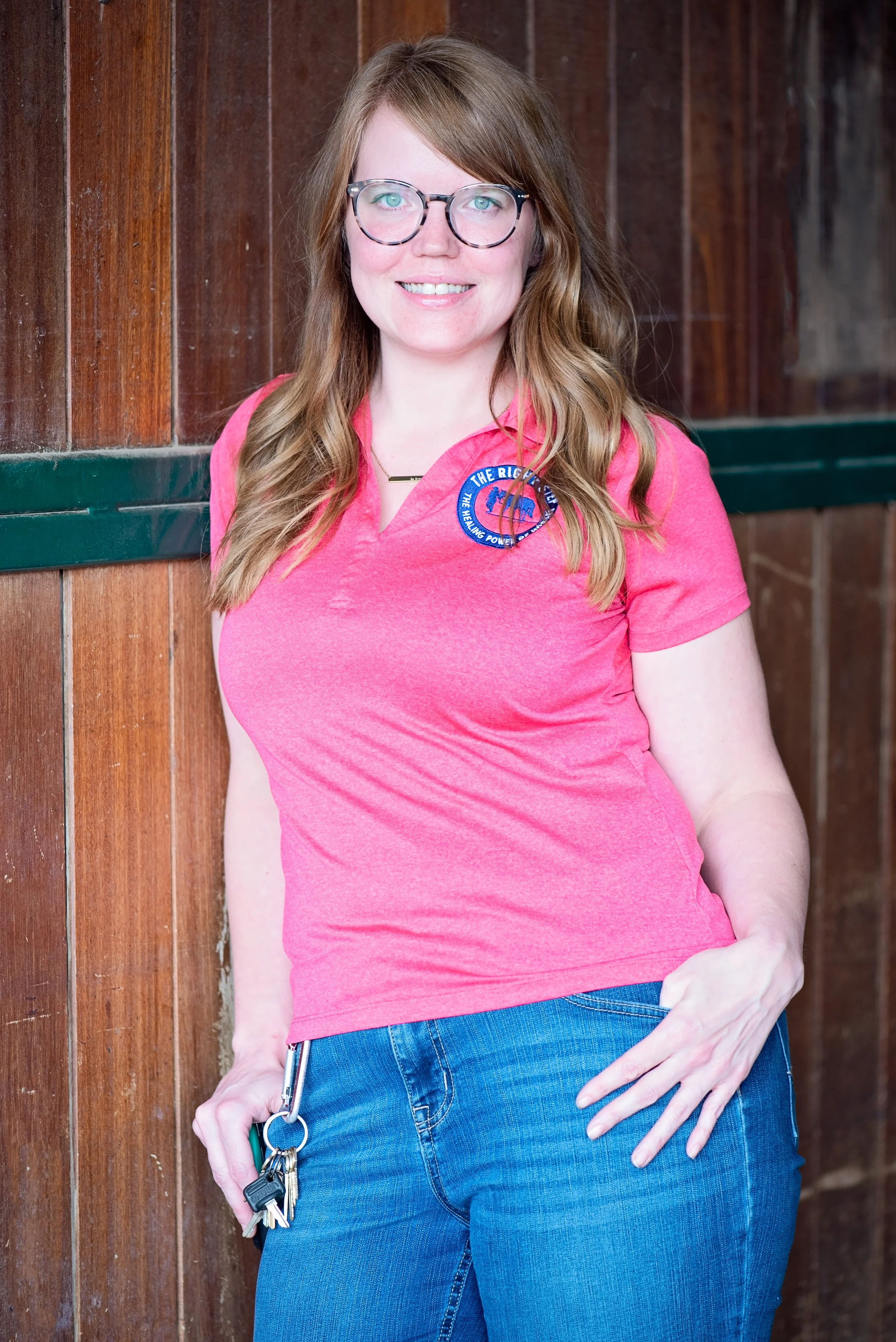 Young woman with long, wavy hair wearing glasses, a pink polo shirt, and blue jeans, smiling in front of a wood-paneled wall.