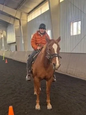 Person riding a horse inside an indoor riding arena, wearing an orange jacket and black helmet, with some cones visible on the ground.