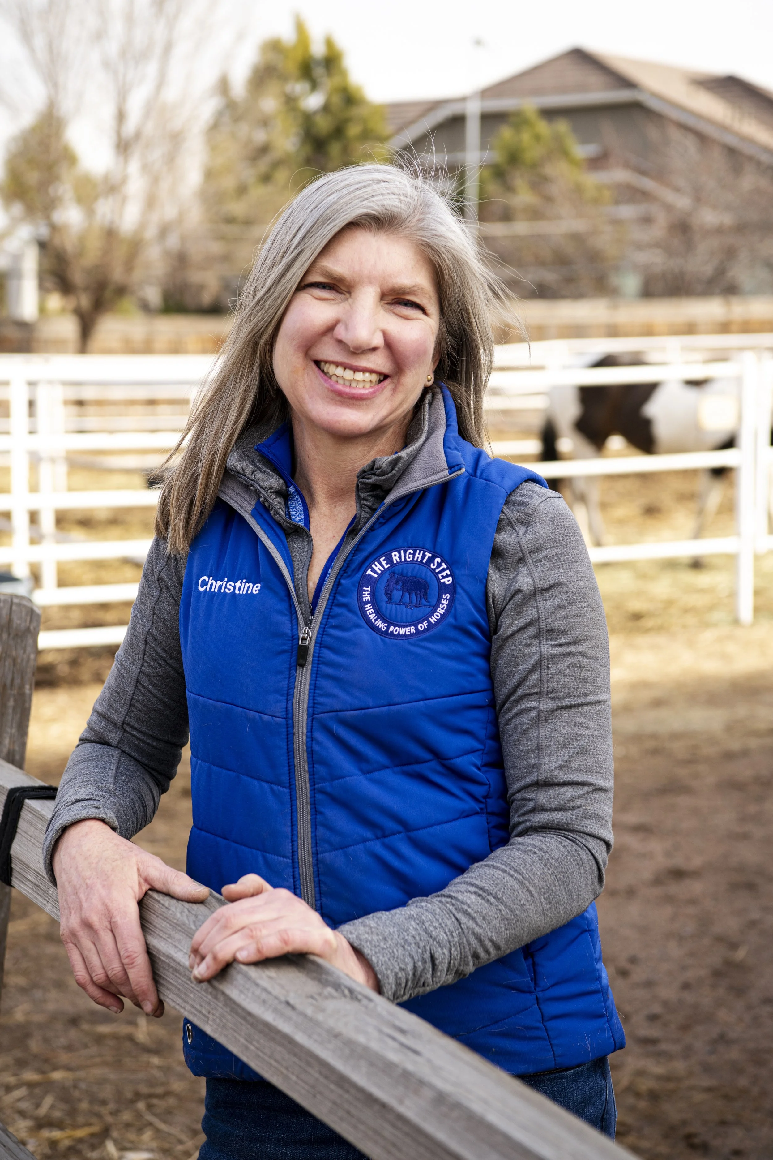 A woman with gray hair smiling outdoors, leaning on a wooden fence at a farm, wearing a blue vest with a logo and the name 'Christine' on it.