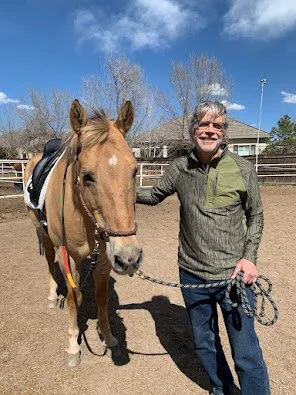 A man standing next to a light brown horse outdoors at a riding arena, holding the horse's reins, on a sunny day with blue skies and some clouds.