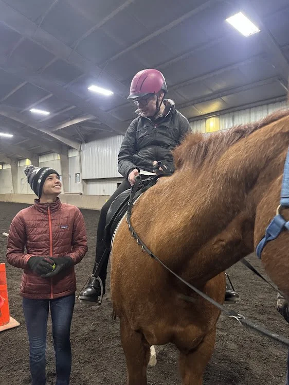 A client is sitting on a brown horse wearing a pink helmet, while a volunteer stands nearby, smiling and dressed in a red jacket and black gloves, inside an indoor riding arena.