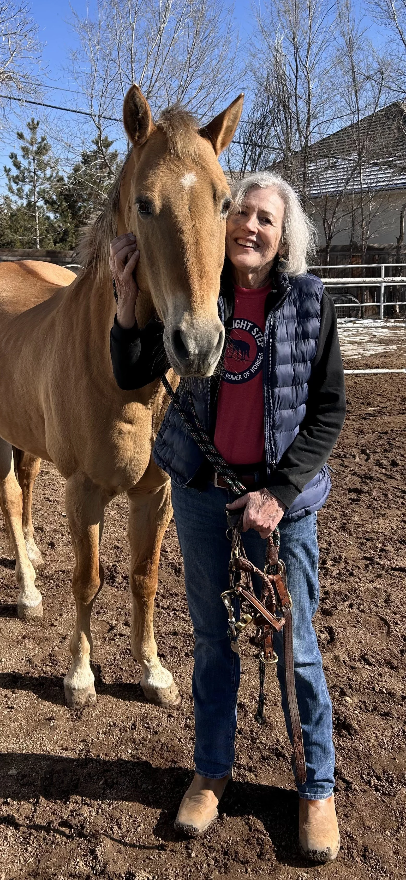 An elderly woman in a black and blue jacket, red shirt, blue jeans, and tan boots smiling while holding a tan horse's halter, standing on a dirt surface with a fenced outdoor area, trees, and a building in the background.