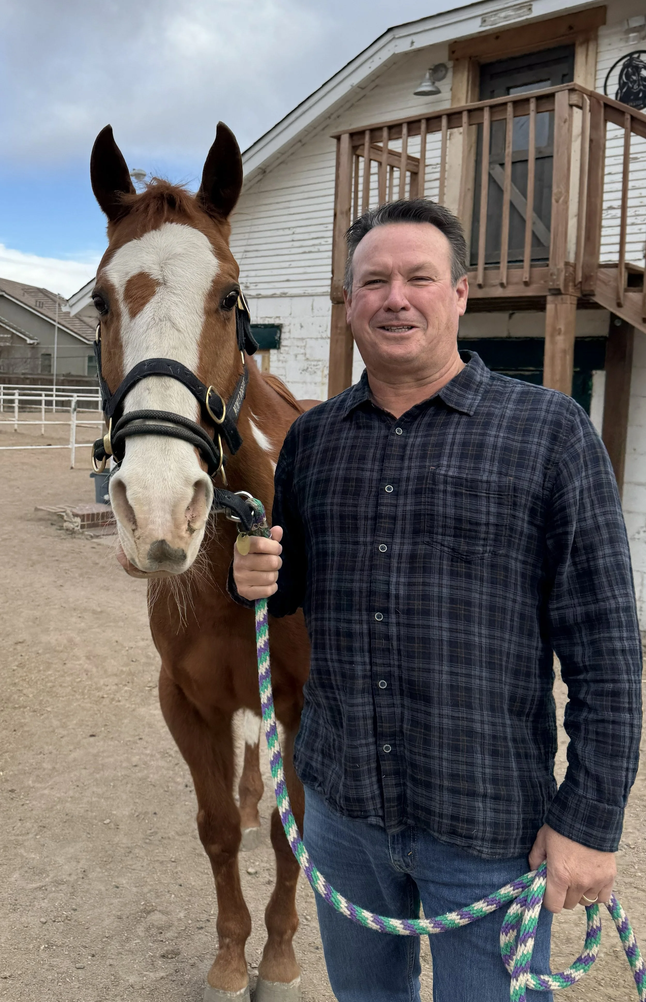 Steve Wright standing outdoors holding the lead of a brown and white horse, with a white brick building with a small wooden balcony in the background.