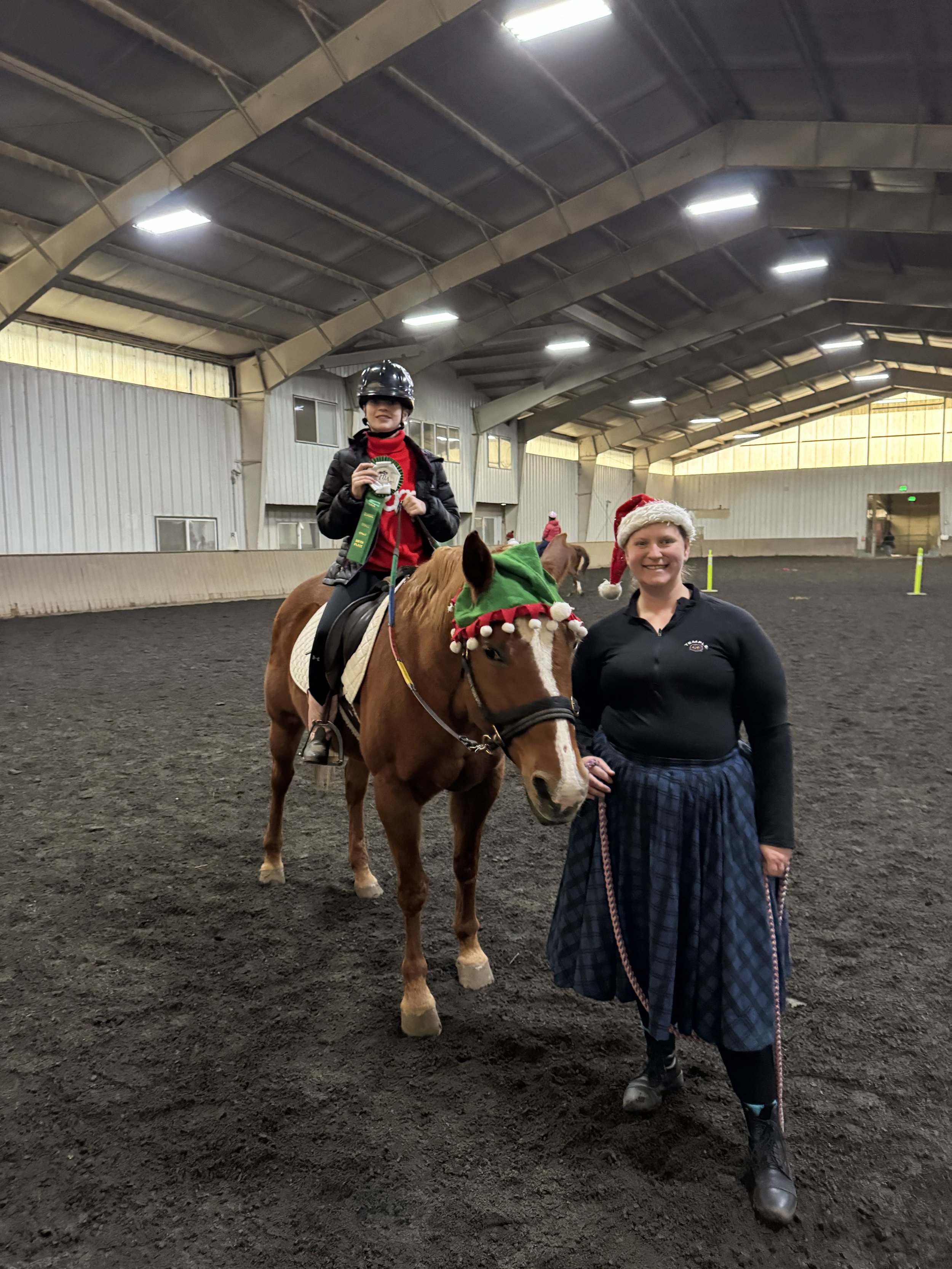 A young girl riding a horse wearing a Santa hat and holding a ribbon, accompanied by a smiling woman holding the horse's lead, inside an indoor equestrian arena.