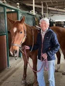 An elderly woman with white hair smiling and holding a chestnut horse with a white blaze on its face inside a stable.