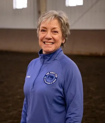Smiling older woman in a blue athletic jacket with a logo, inside a large indoor sports facility.