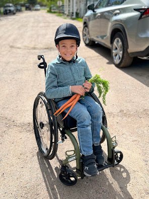 A young The Right Step Inc. client in a wheelchair wearing a helmet, holding carrots and a bunch of greens, outdoors on a dirt path with cars parked in the background.