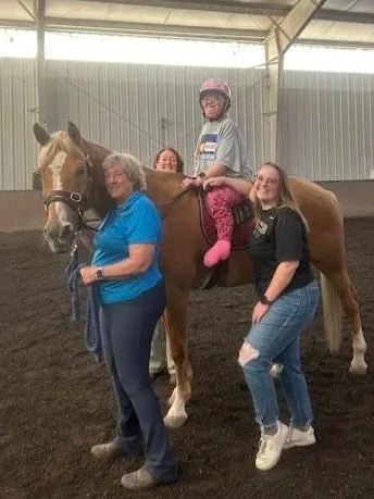 A young girl wearing a helmet riding a brown horse with a white blaze, accompanied by three women standing nearby inside a covered riding arena.