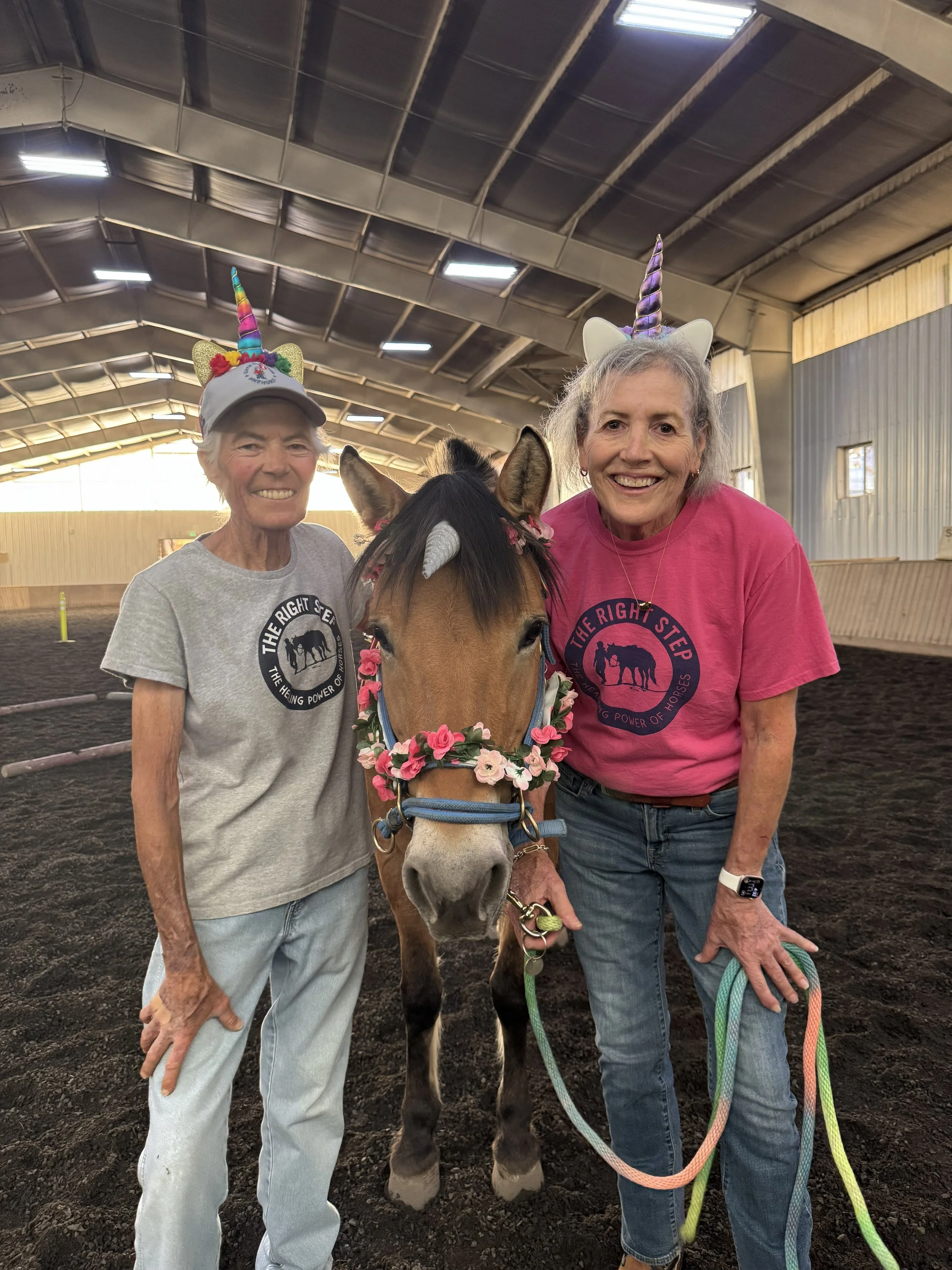 Two women and a decorated horse inside an indoor riding arena. The women wear unicorn headbands and TRS shirts, smiling at the camera.