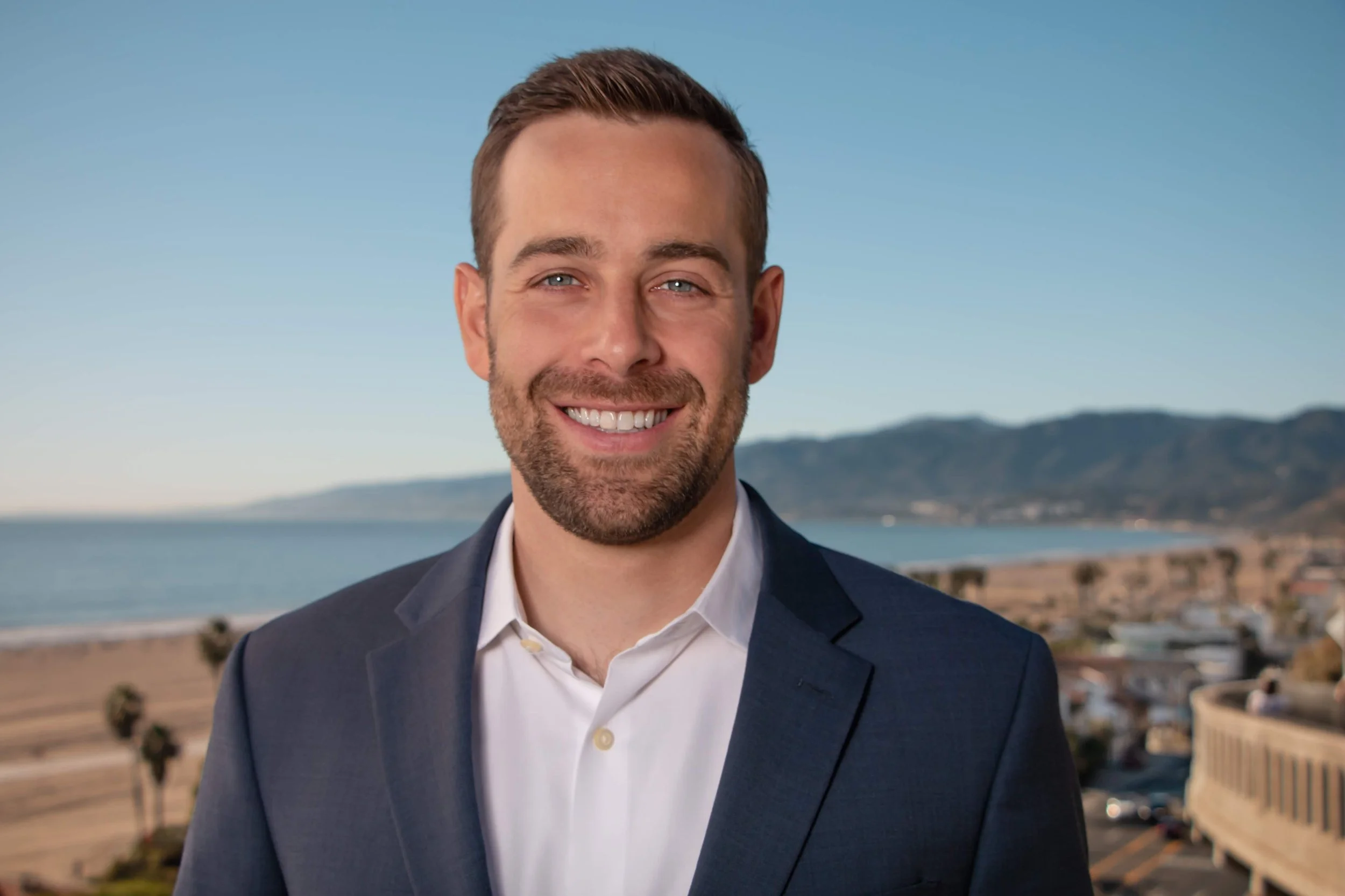 Headshot of Cory Shumaker in suit in front of coastline