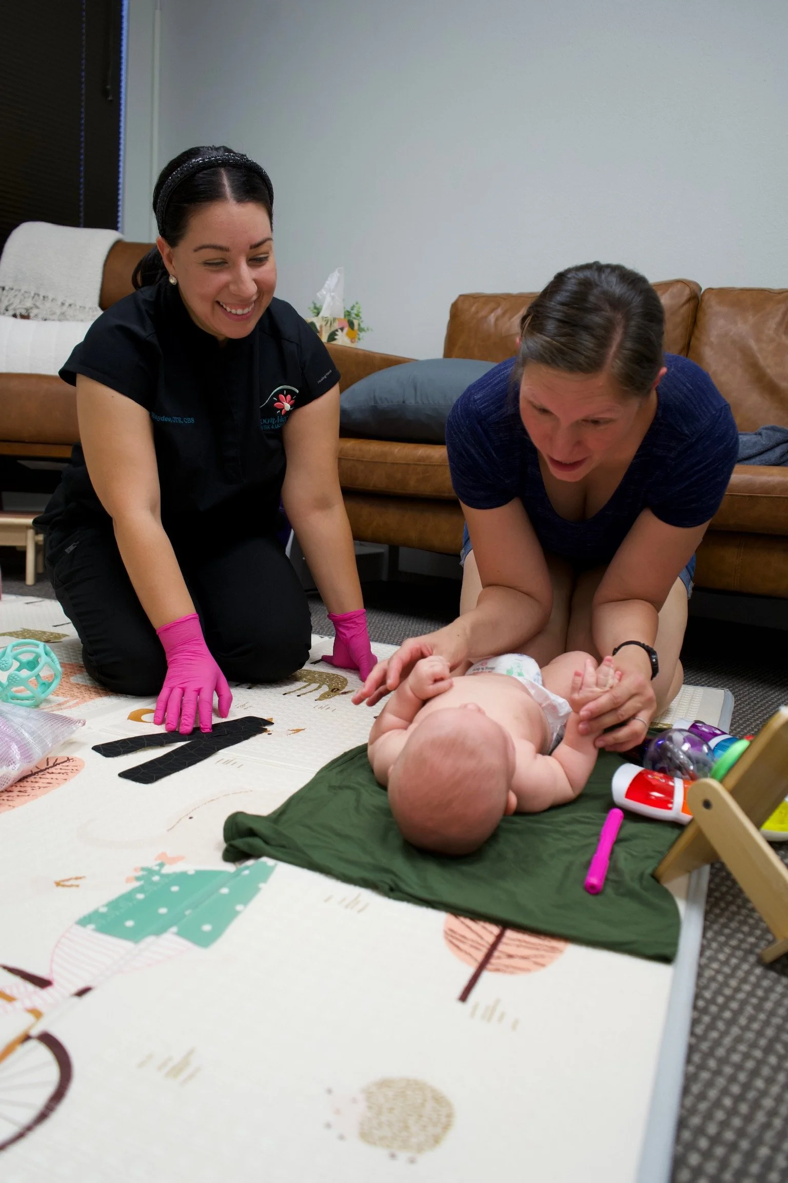 Haydee Medrano kneeling next to a mom and baby. Mom is also kneeling and the baby is laying on their back looking at the mom.