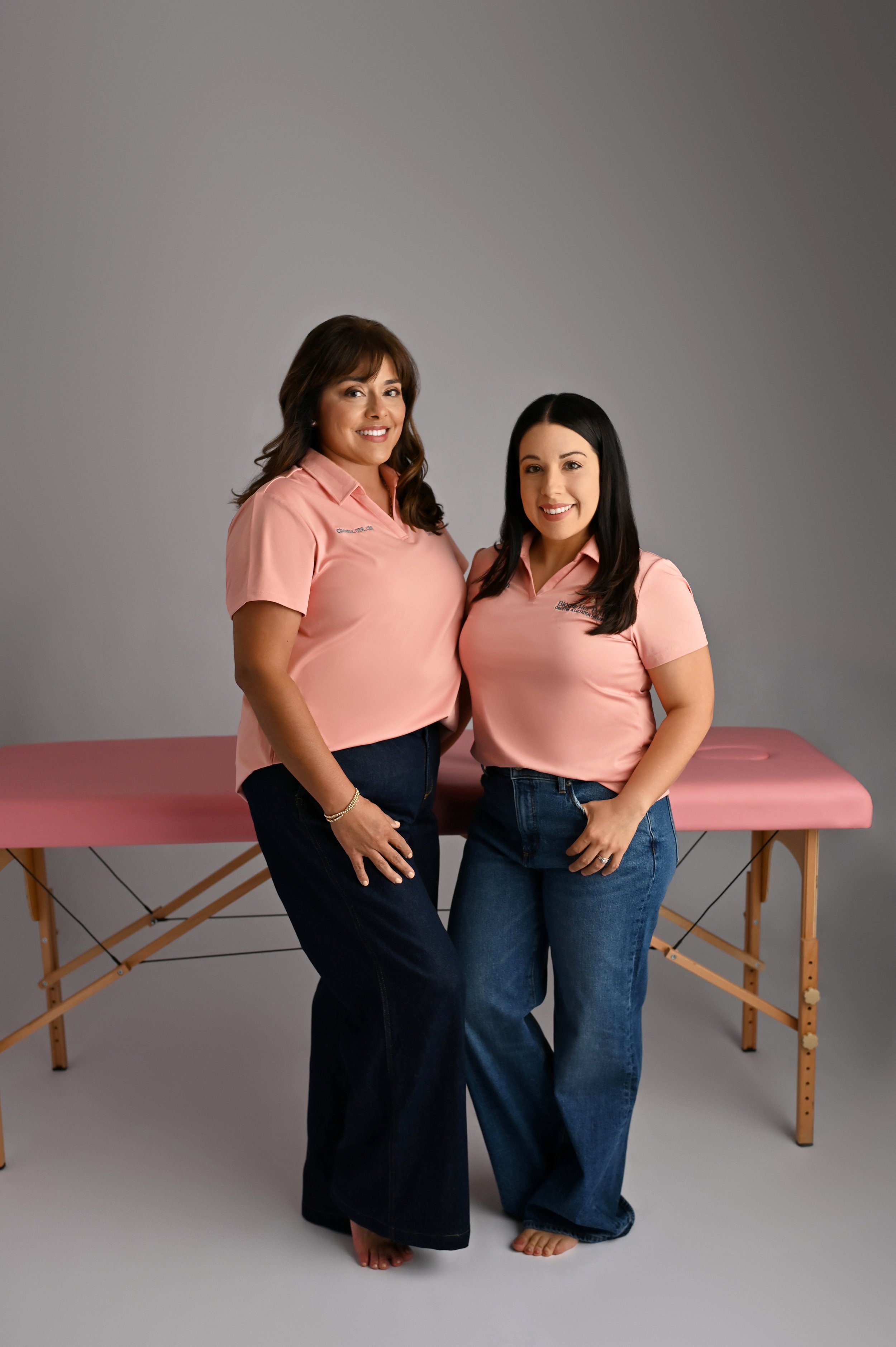 Christine Hernandez and Haydee Medrano pose in pink polo shirts in front of a mat table at Bloom Her Path in Corpus Christi, TX where they provide pelvic floor therapy for women