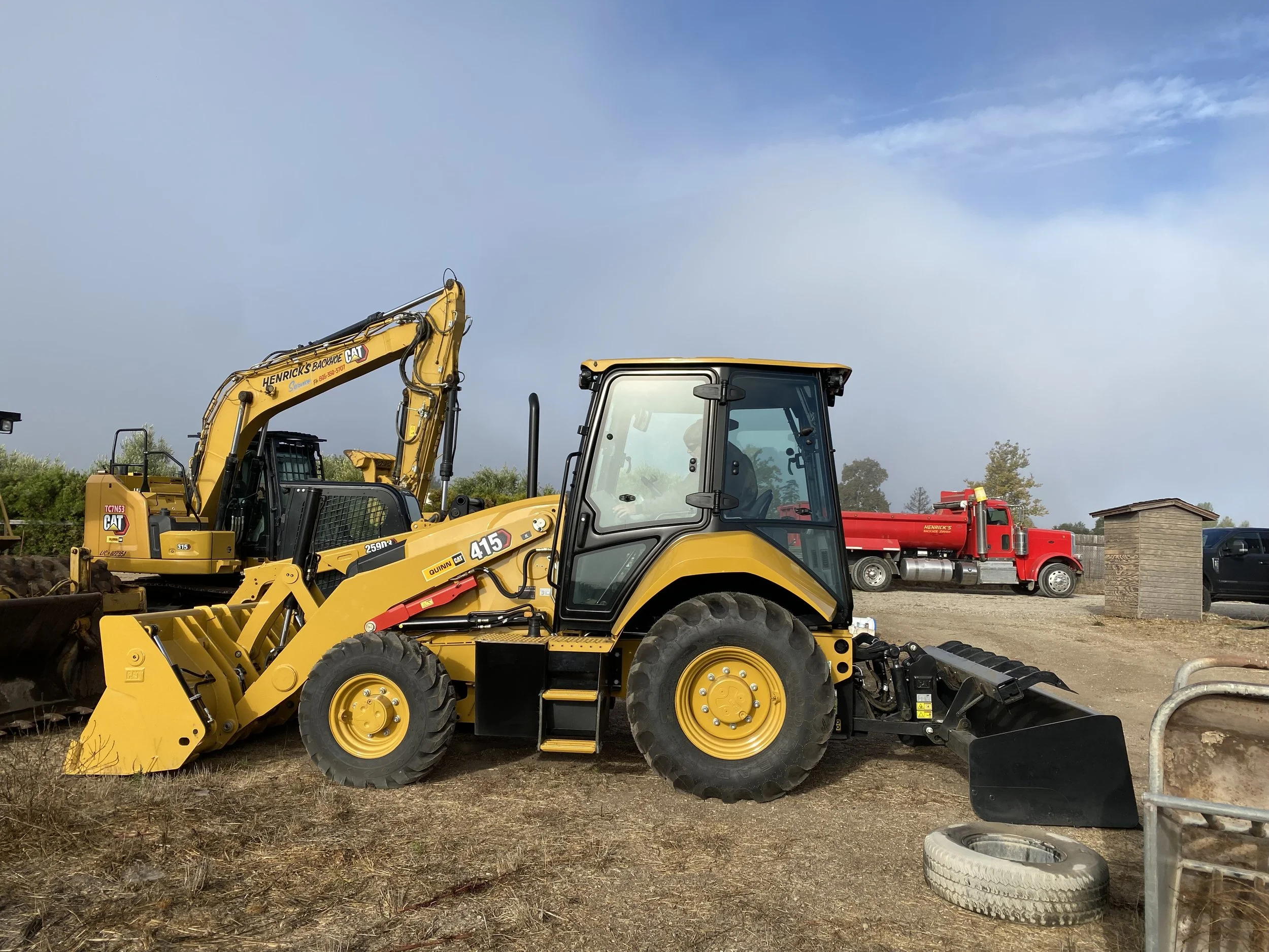 A small excavator on a construction site digging into the dirt with houses and trees in the background.