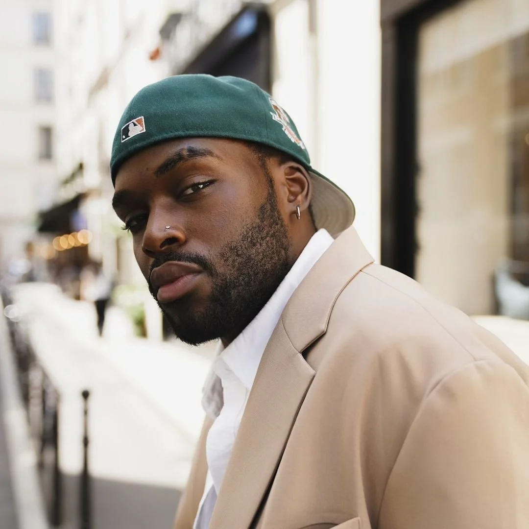 A young man with a beard, wearing a white shirt, beige blazer, green baseball cap tilted backward, and a nose piercing, standing on a city street with blurred background buildings.