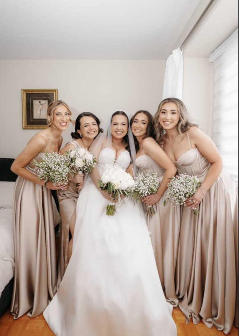 Bride in a white wedding dress surrounded by five bridesmaids in matching satin dresses, all holding bouquets of white flowers, standing in a room with white walls and a window with sheer curtains.