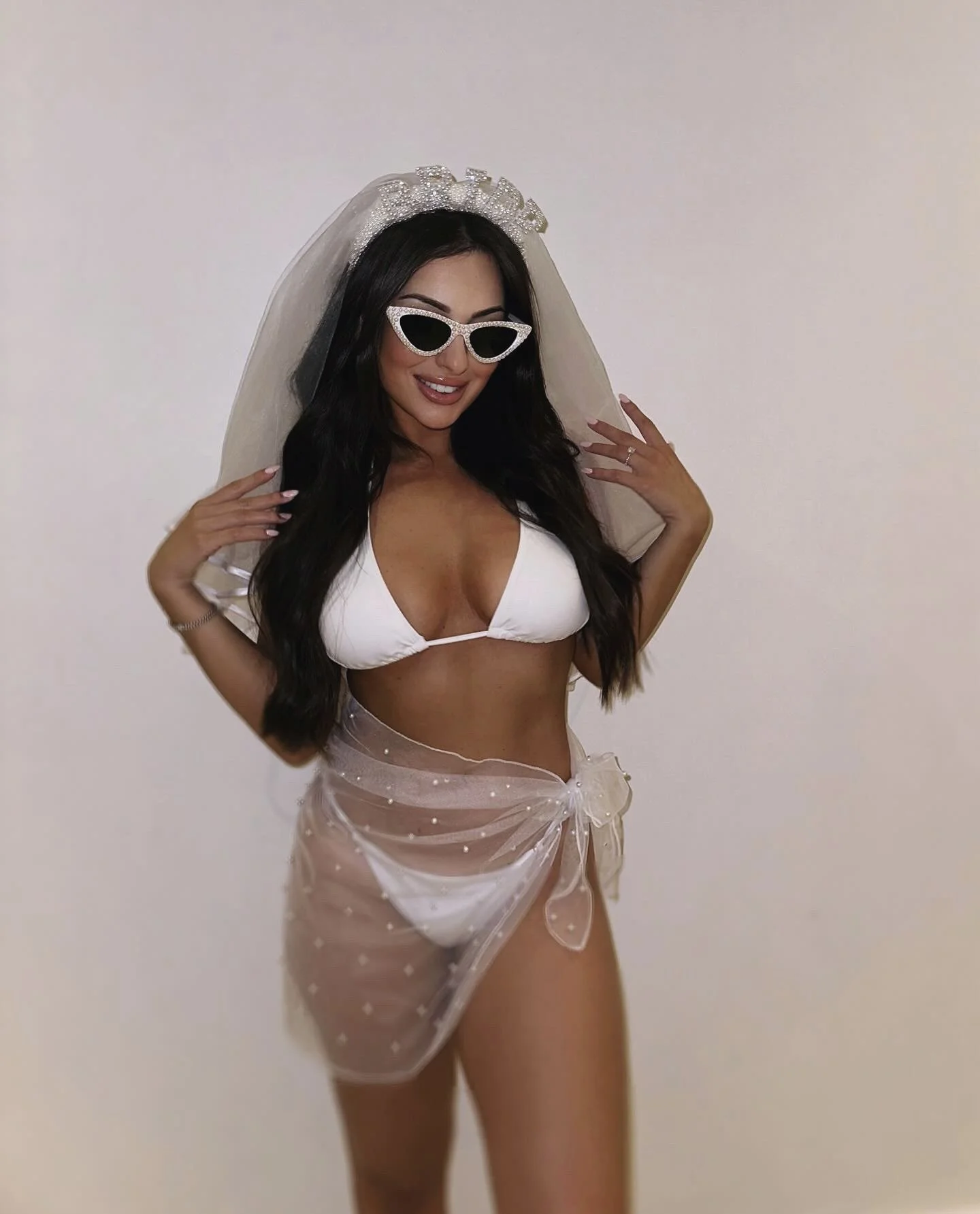 Woman in a bikini with a sheer polka dot cover-up, wearing sunglasses and a veil, posing with a smile against a plain background