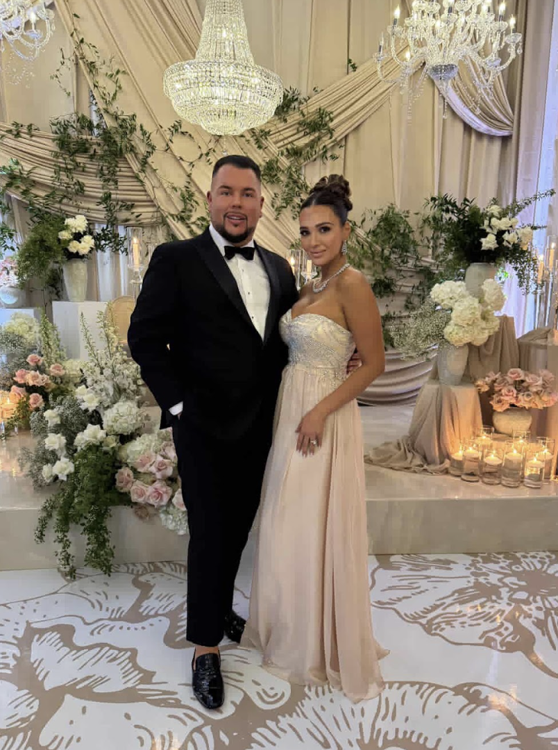 A man in a black tuxedo and bow tie standing next to a woman in an elegant strapless cream-colored gown, at a decorated wedding or formal event with floral arrangements, chandeliers, and candles.