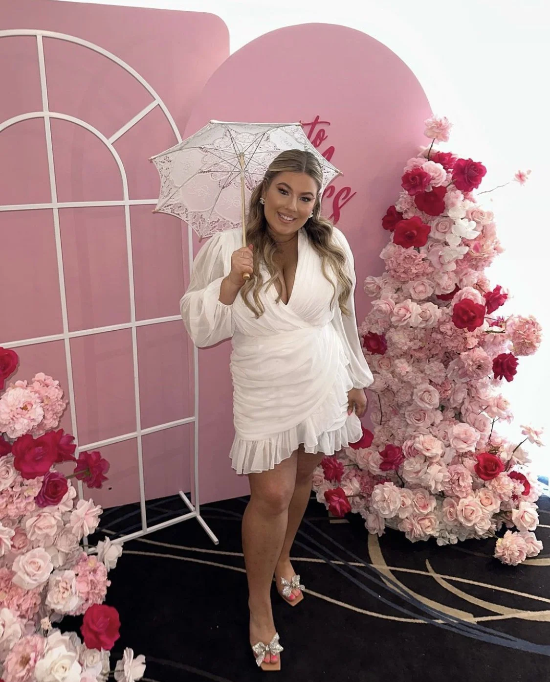 A woman in a white dress holding a lace parasol posing in front of a pink backdrop with flowers.