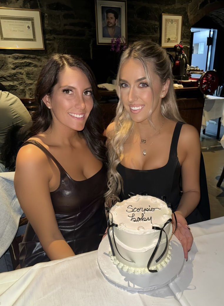 Two women sitting at a table celebrating with a cake labeled "Scorpio Ladies". The woman on the left has dark hair and is wearing a black leather dress. The woman on the right has blonde hair with loose waves and is wearing a black dress with thick s