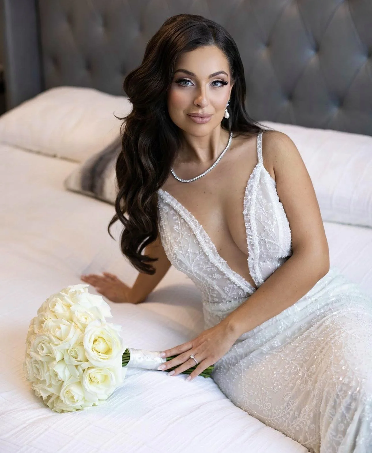 A bride with long dark hair and blue eyes wearing a white wedding dress with beaded details, sitting on a bed with a bouquet of white roses.