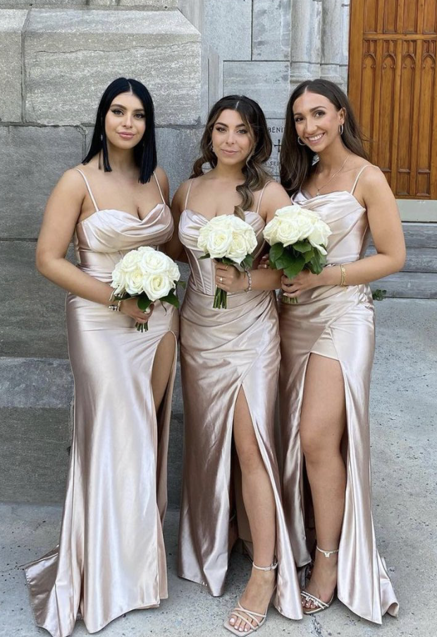 Three women in satin gowns holding white bouquets, standing outdoors against a stone wall with a wooden door in the background.