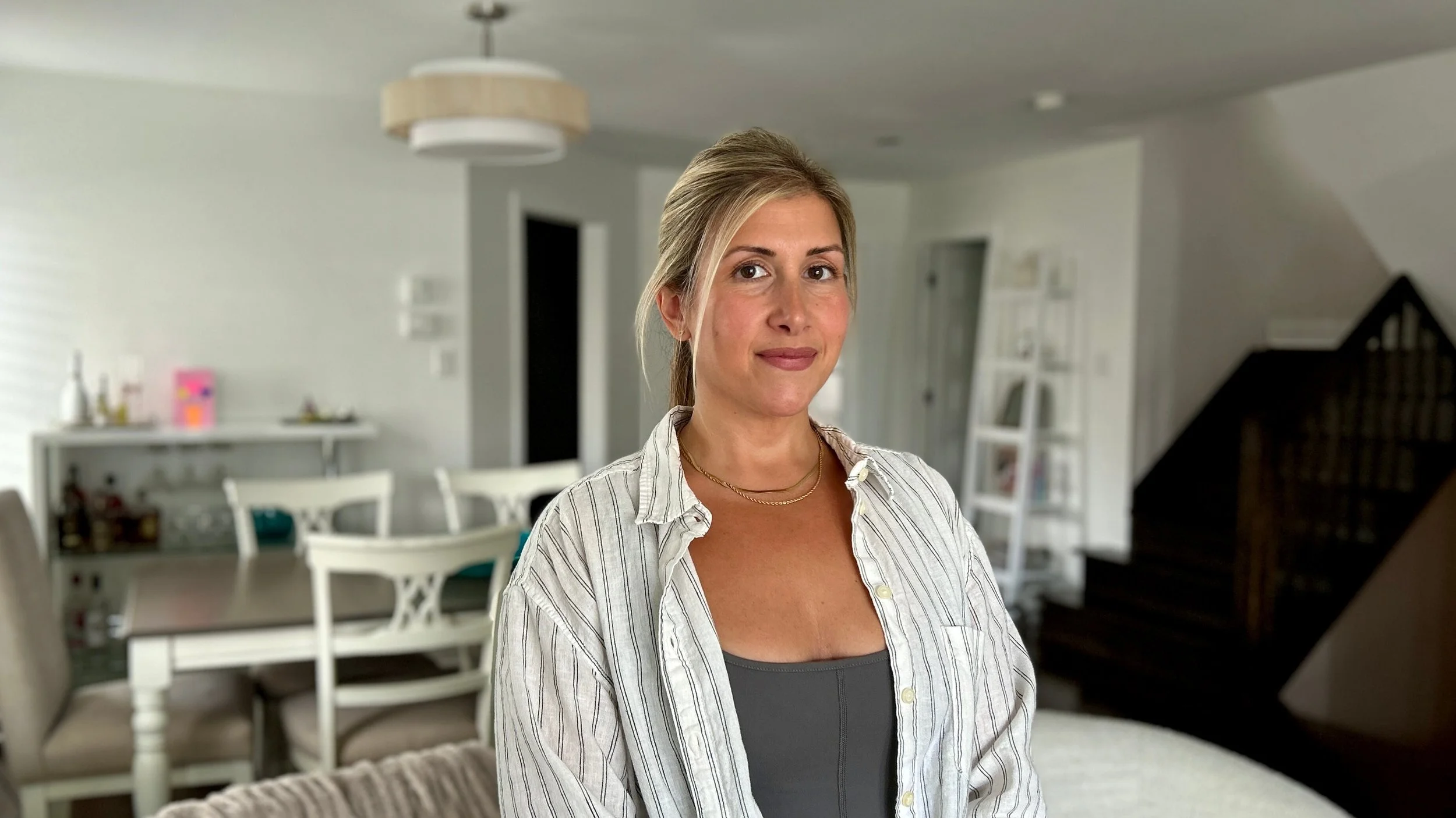 A woman with blonde hair in a ponytail, wearing a striped button-up shirt over a gray top, standing in a modern, well-lit living or dining area with white walls and furniture.