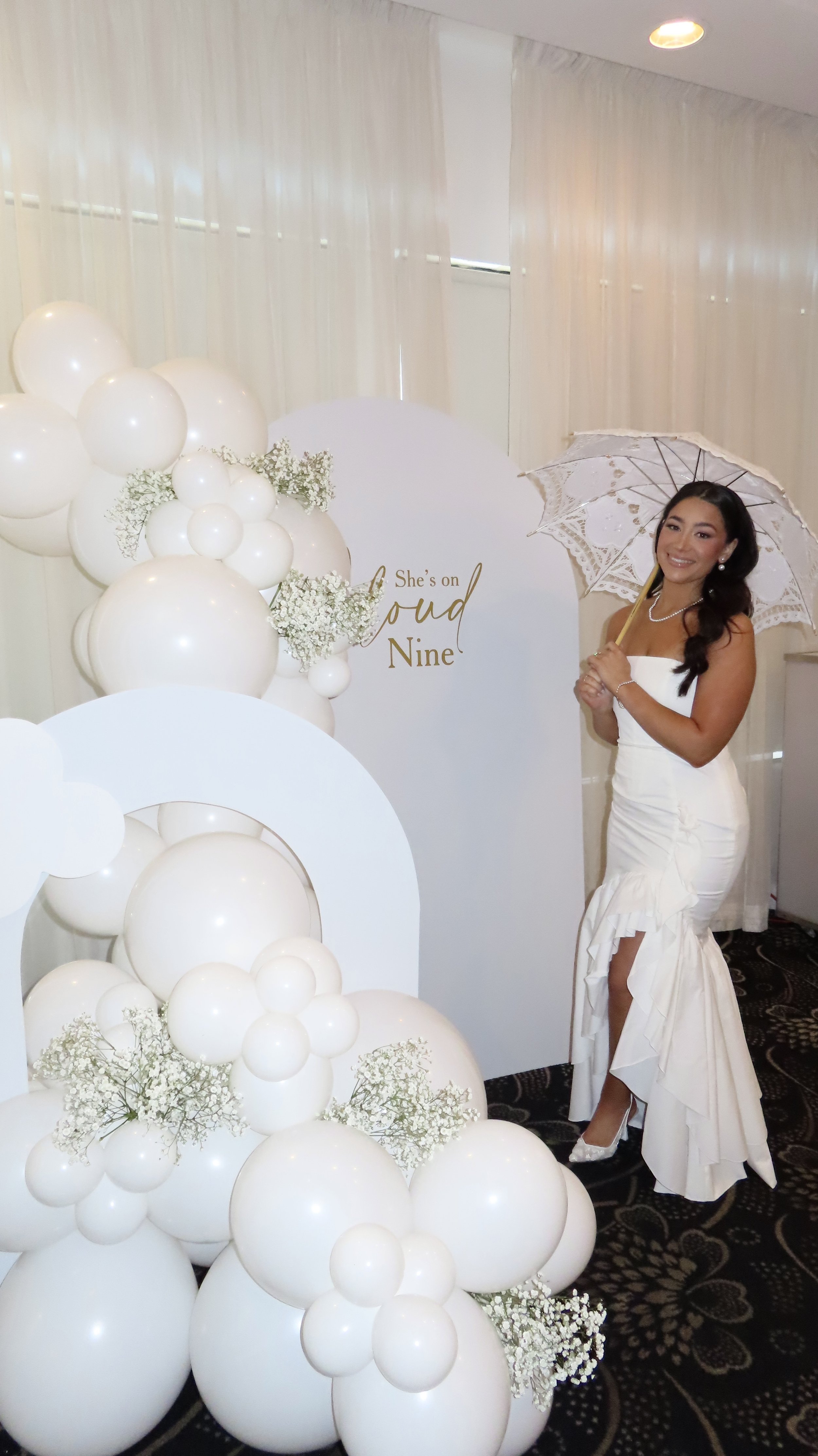 A woman in a white wedding dress holding a lace parasol, standing next to a balloon and floral decoration with a sign that reads "She's on Cloud Nine."
