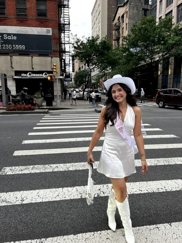A young woman dressed in a white dress, white cowboy hat, and white boots, smiling and walking across a crosswalk in an urban city street.