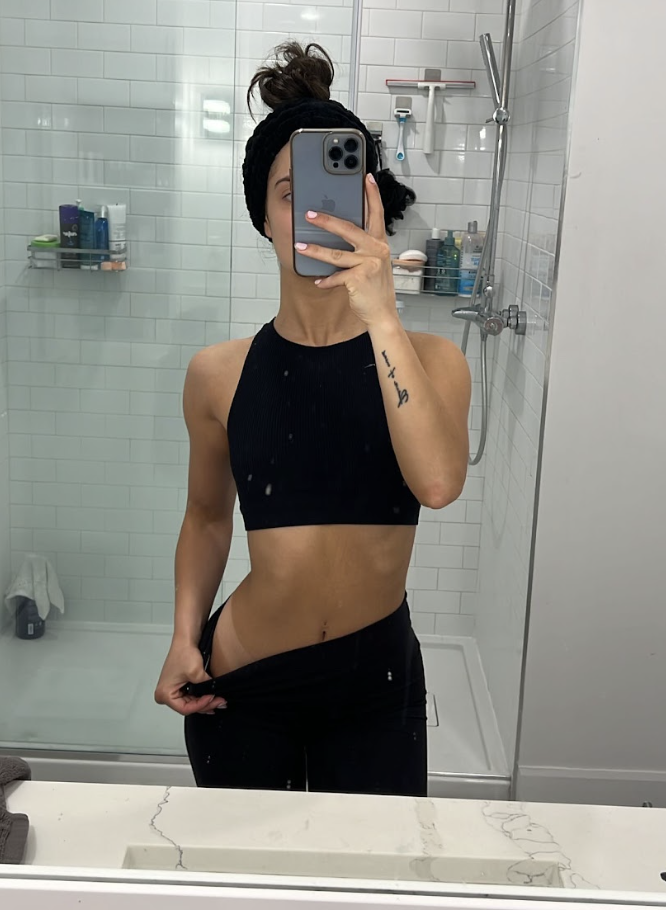 Woman taking a mirror selfie in a bathroom, wearing a black sports bra and black pants, with her hair in a messy bun.