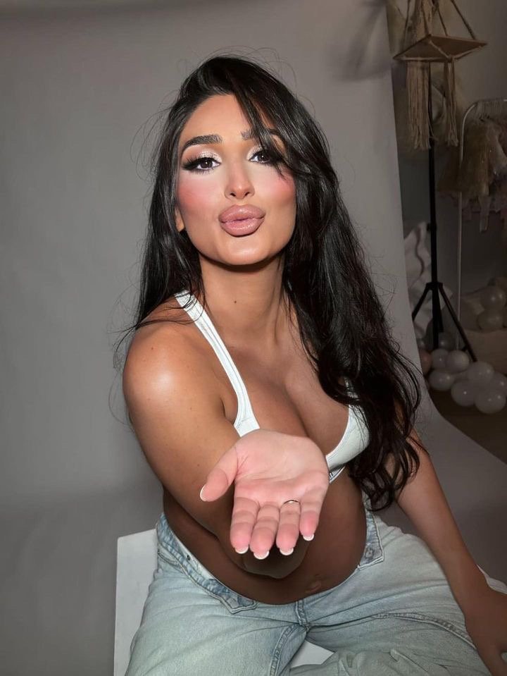 Young woman with long dark hair and makeup, sitting down in a white tank top and light jeans, extending her hand towards the camera, inside a well-lit studio.