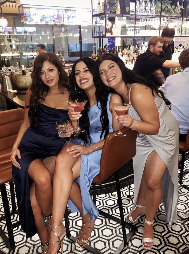 Three women sitting at a bar, smiling and holding cocktails, dressed in elegant evening wear in a lively bar or restaurant.