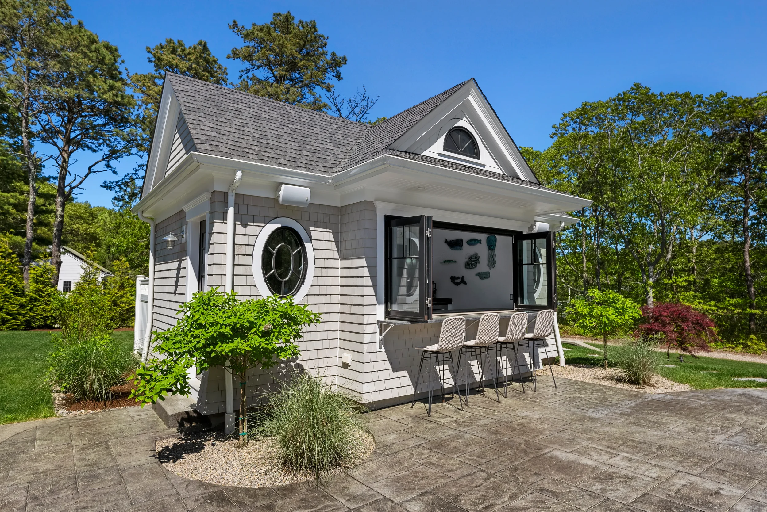 An outdoor view of a small white house with a gray shingled roof, black window frames, and an open window with a bar and four beige chairs. There's a garden with trees and shrubs, and a patio with a stamped concrete surface.