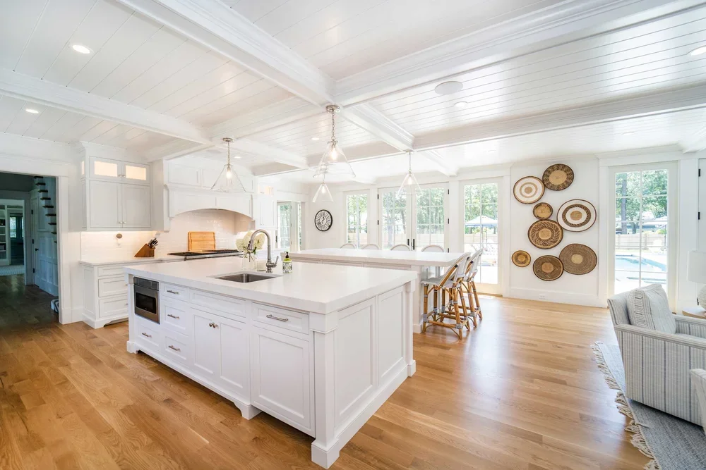 Bright, white kitchen with a large island, wooden floors, and a dining area with barstools. Natural light from large windows, modern decor, and wall art.