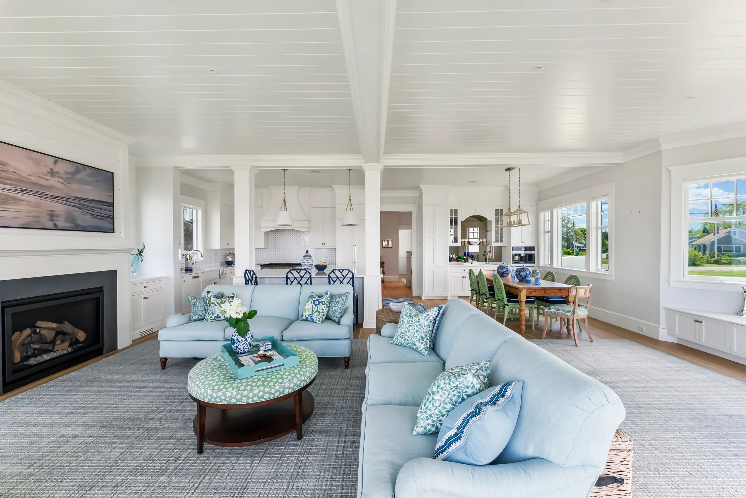 Bright and spacious living room with white walls, large windows, and light blue sofas decorated with patterned pillows. A fireplace is visible on the left and a wooden dining table with green chairs is in the background. The room has a coastal decor
