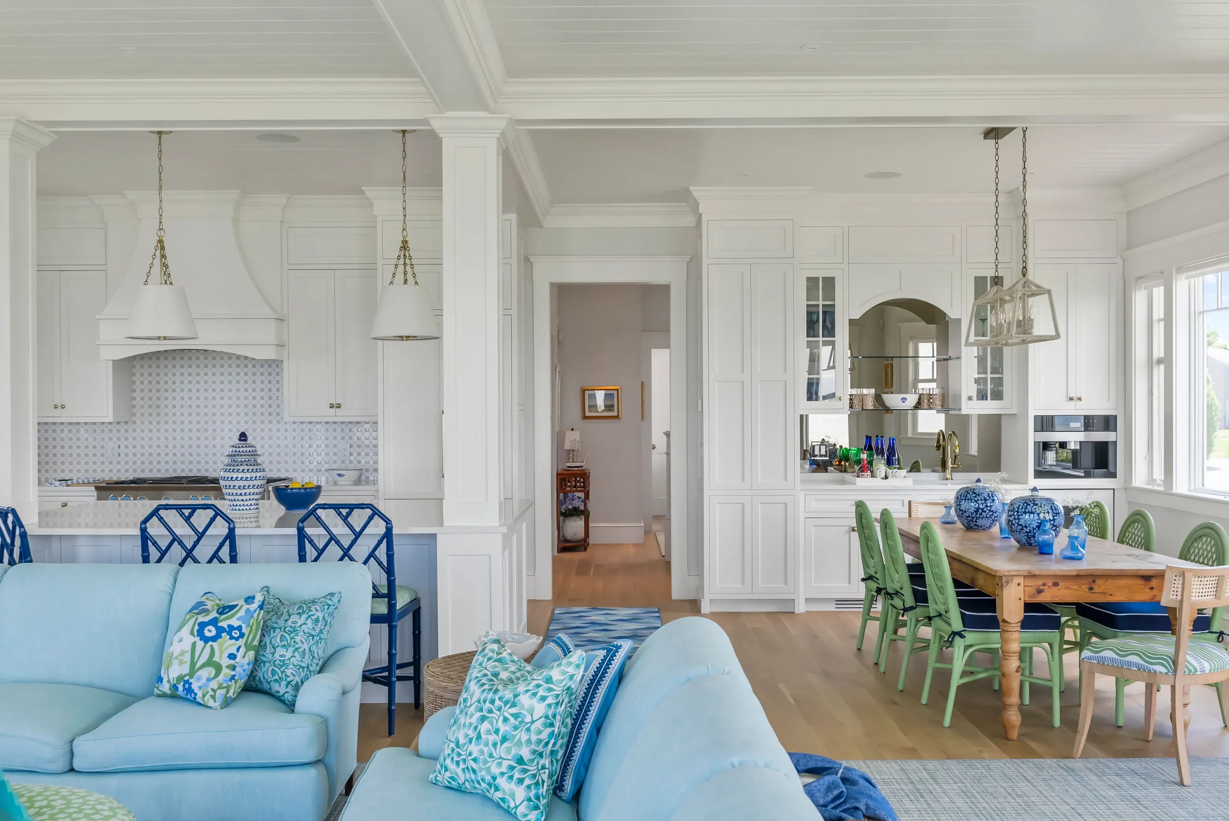 Bright open-concept kitchen and dining area in a house, with white cabinets, large windows, a wooden dining table with green chairs, blue decorative vases, and light wood flooring.