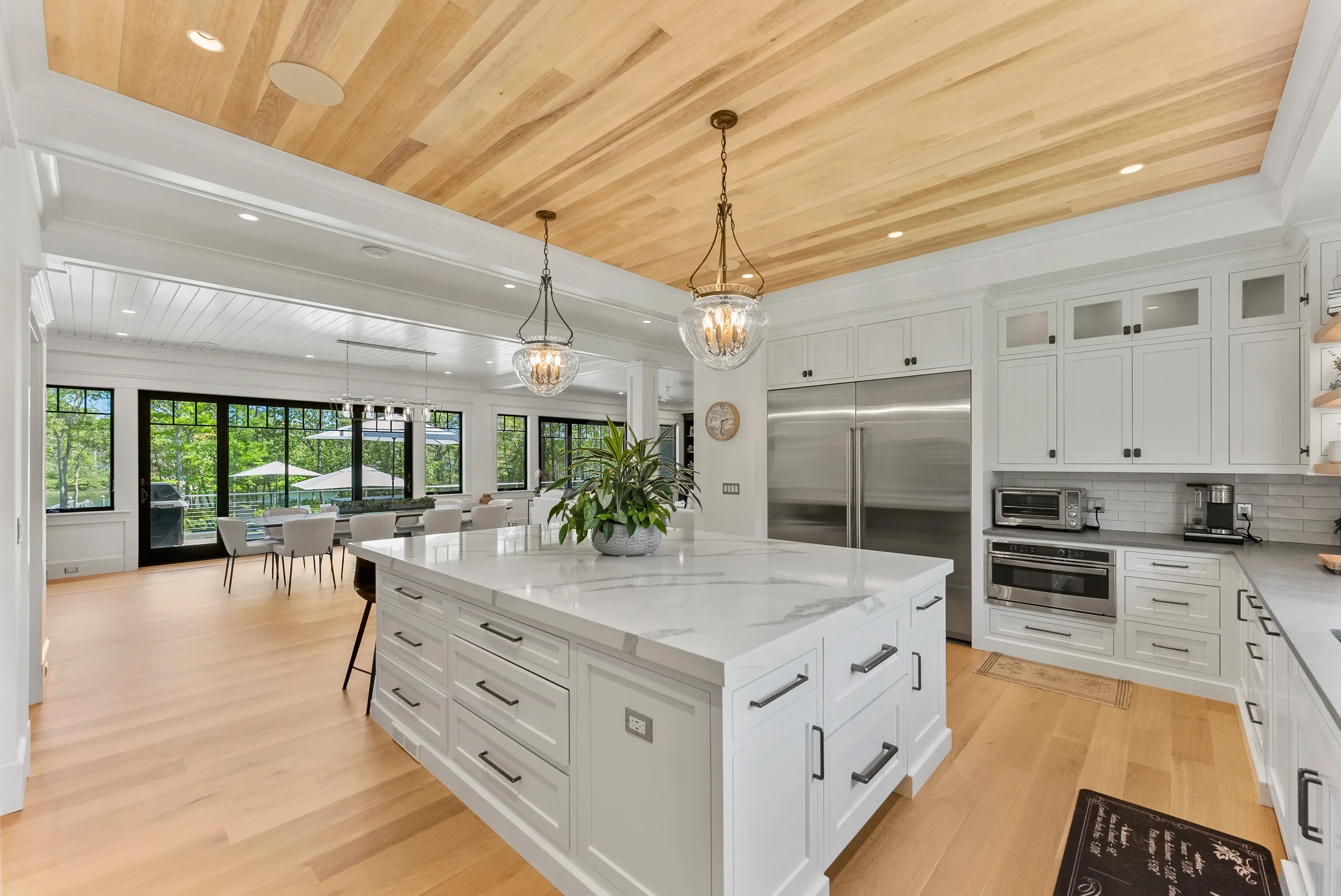 Modern kitchen with white cabinetry and marble countertops, stainless steel appliances, a central island with a plant, hanging pendant lights, and a dining area with large windows and outdoor views.