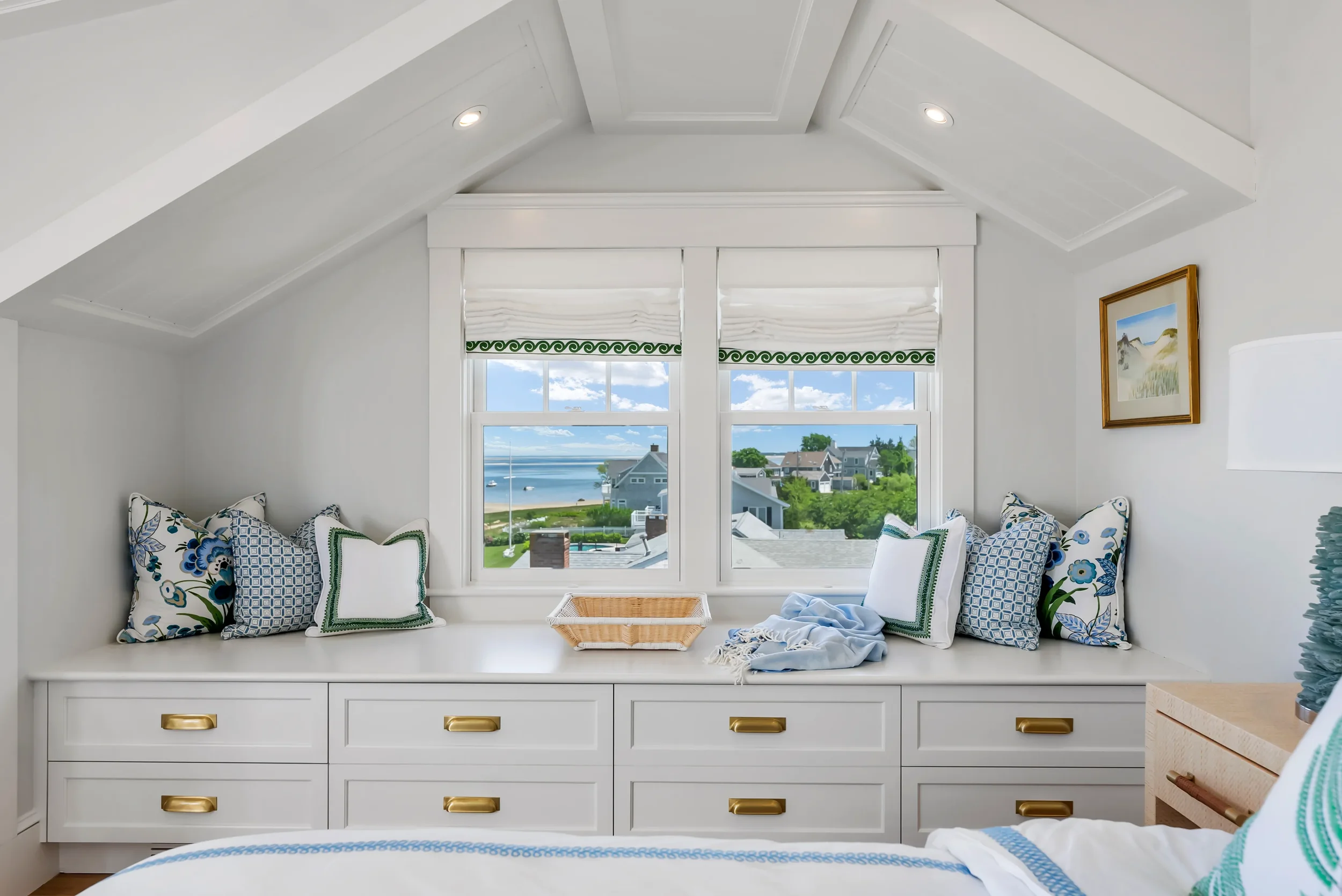A cozy bedroom with a built-in window seat area under a large window, overlooking a beach and houses outside. The window seat has several decorative pillows, and the room is decorated in light, coastal colors with white furniture and accents.