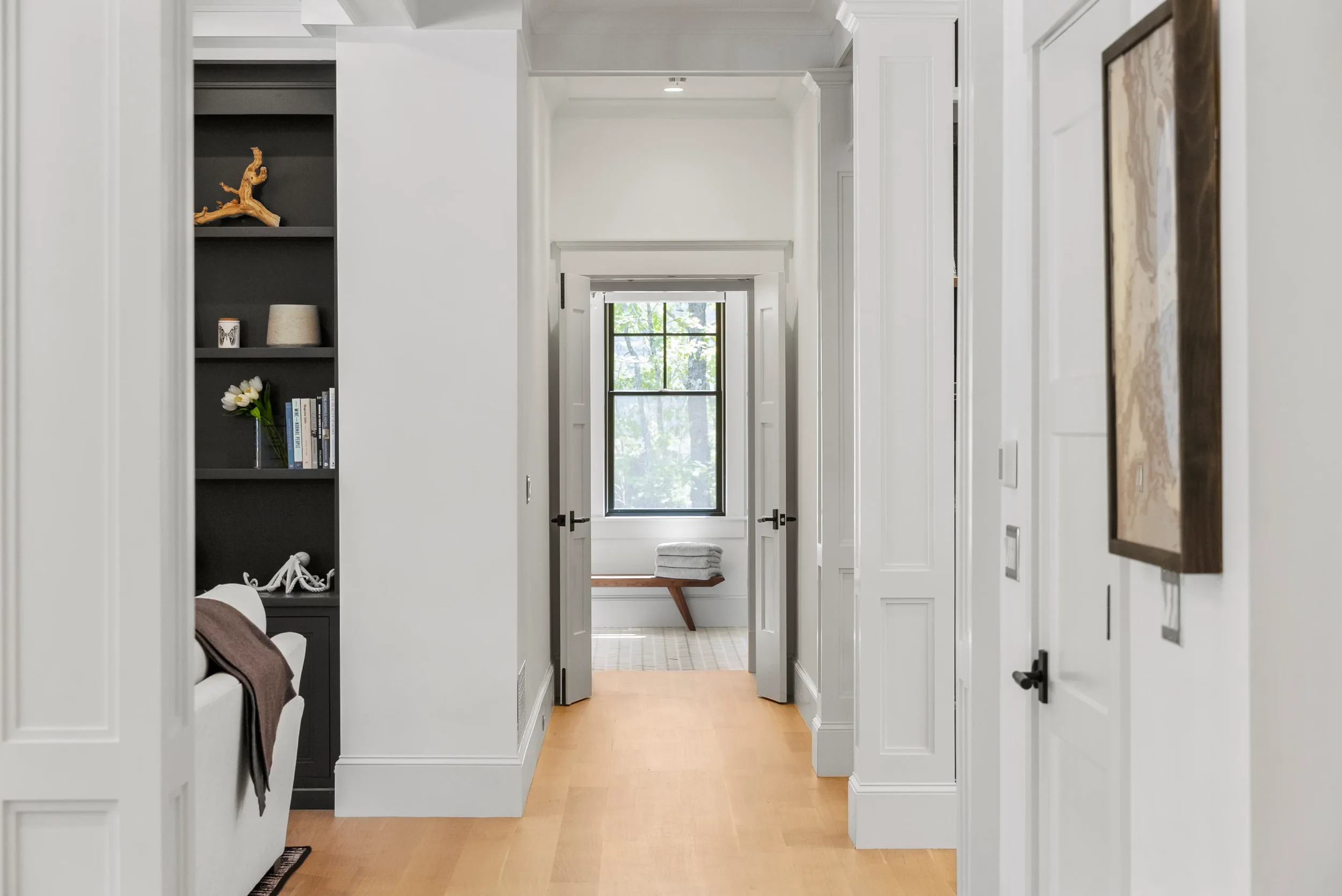 A bright hallway with white walls and trim, leading to a room with a window, a bench with folded towels, and a view of trees outside.