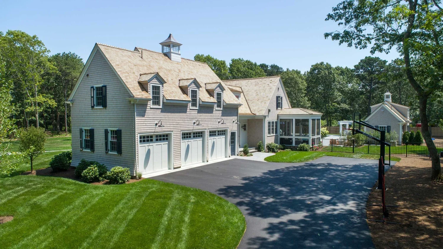 A large house with a three-car garage, a porch, and a basketball hoop in the driveway, surrounded by lush green lawn and trees.