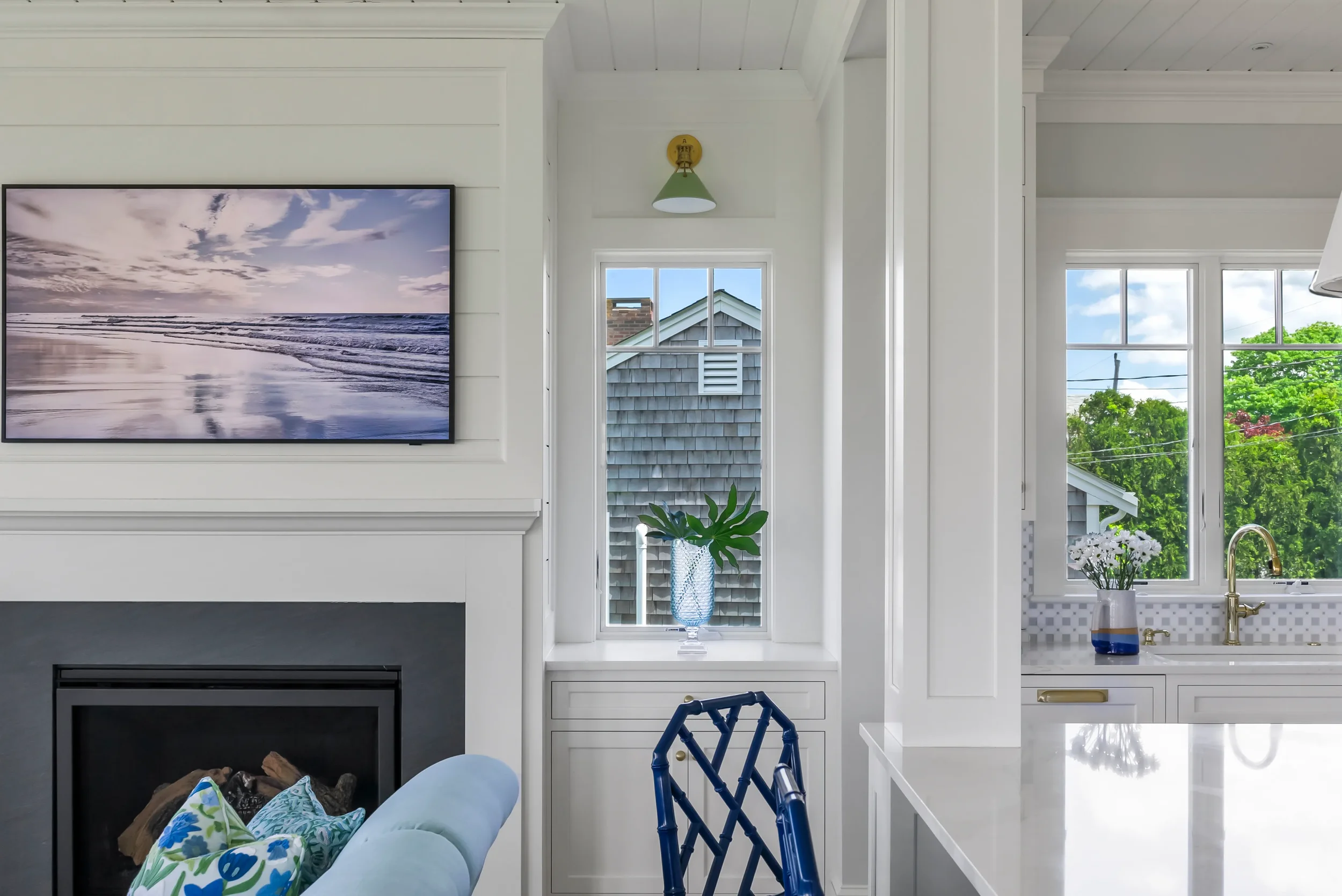 Interior view of a bright kitchen and living room with a window showing green trees, a vase with flowers, and a television on a white fireplace mantel.