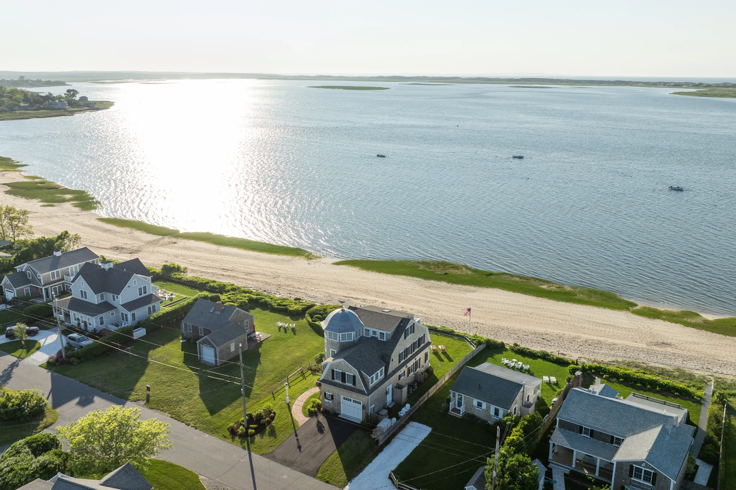 Aerial view of a coastal neighborhood with houses, yards, and a sandy beach along a large body of water, boats on the water, and a bright sunlight reflection on the water surface.