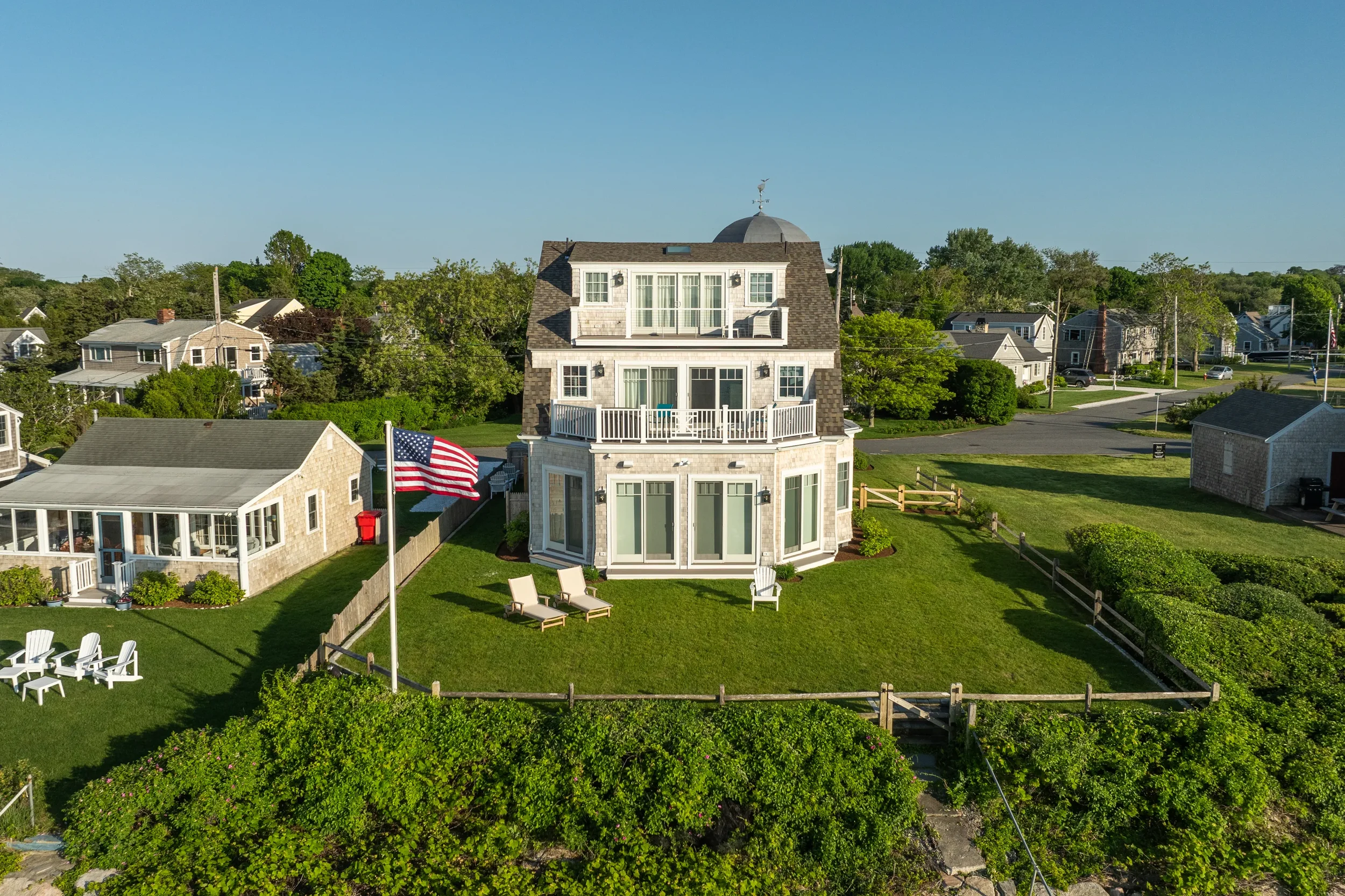 A large multi-story house with white walls and black roofing, with balconies and large windows, surrounded by a lush green yard with outdoor chairs and an American flag on a flagpole.