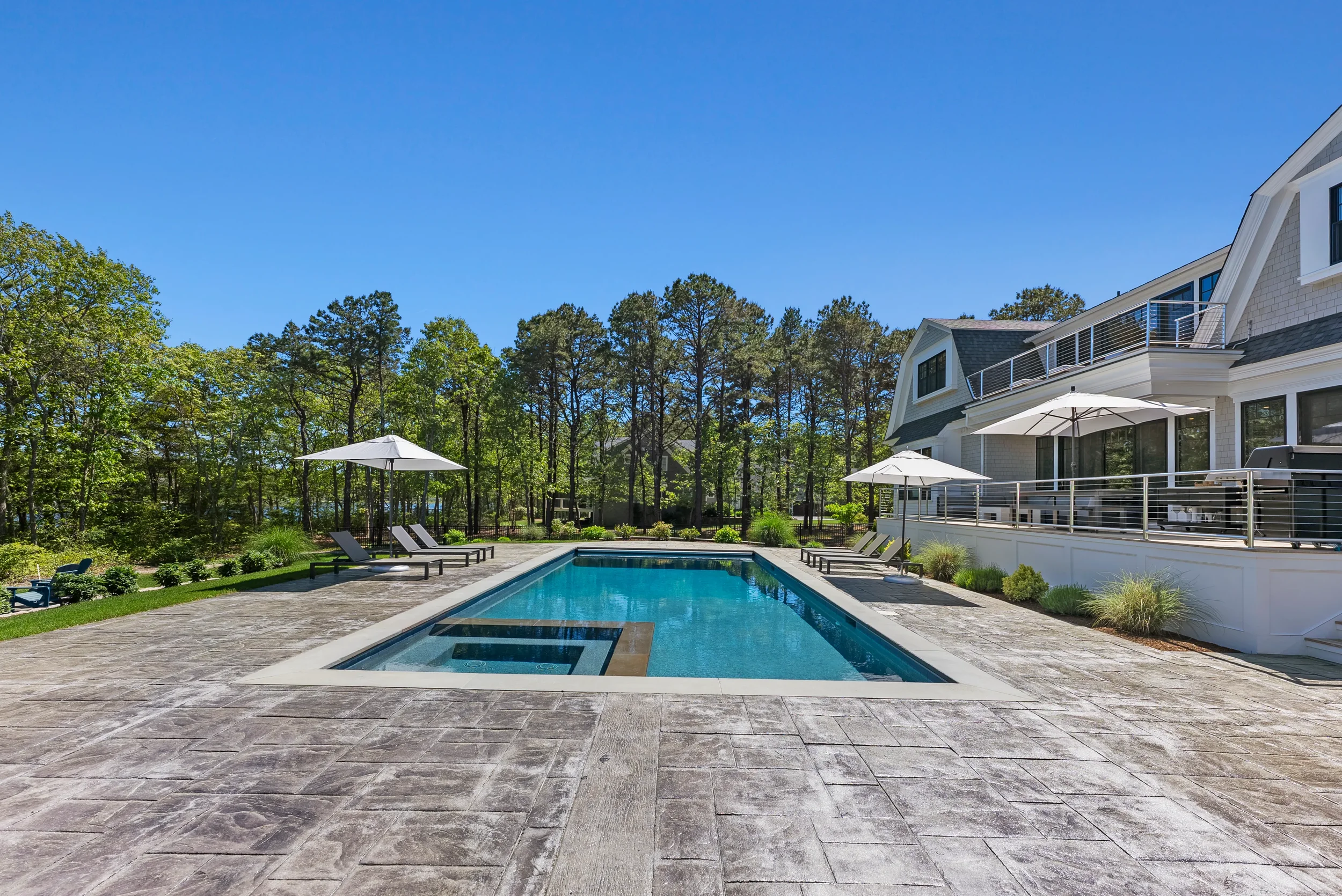 Clean outdoor swimming pool with lounge chairs and umbrellas on a bright sunny day, behind a house with a deck and surrounded by trees.