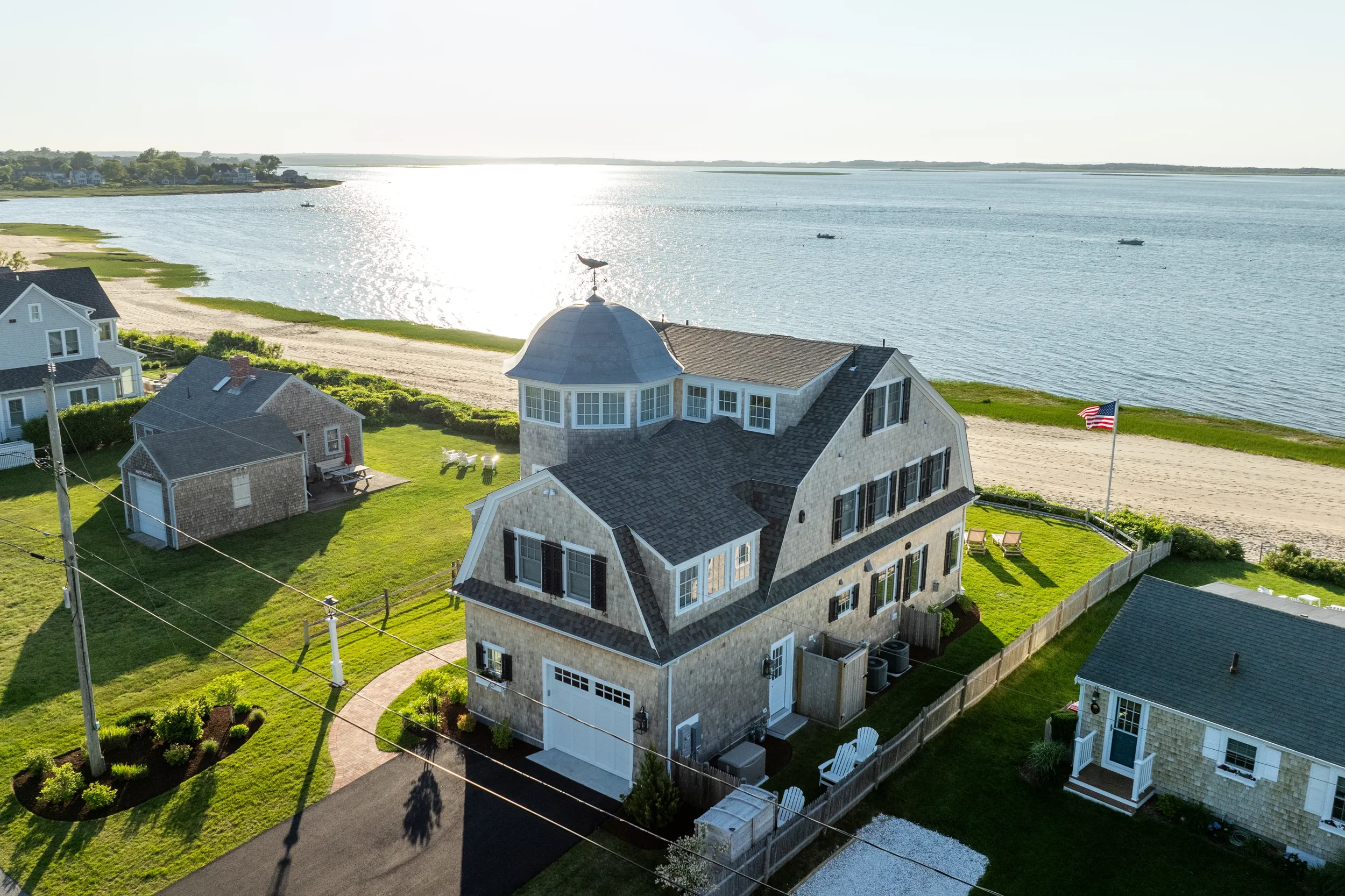 Aerial view of a beach house with a slate roof, windows, and a small tower with a weather vane, located along a sandy beach with water and boats, American flag, and neighboring houses.
