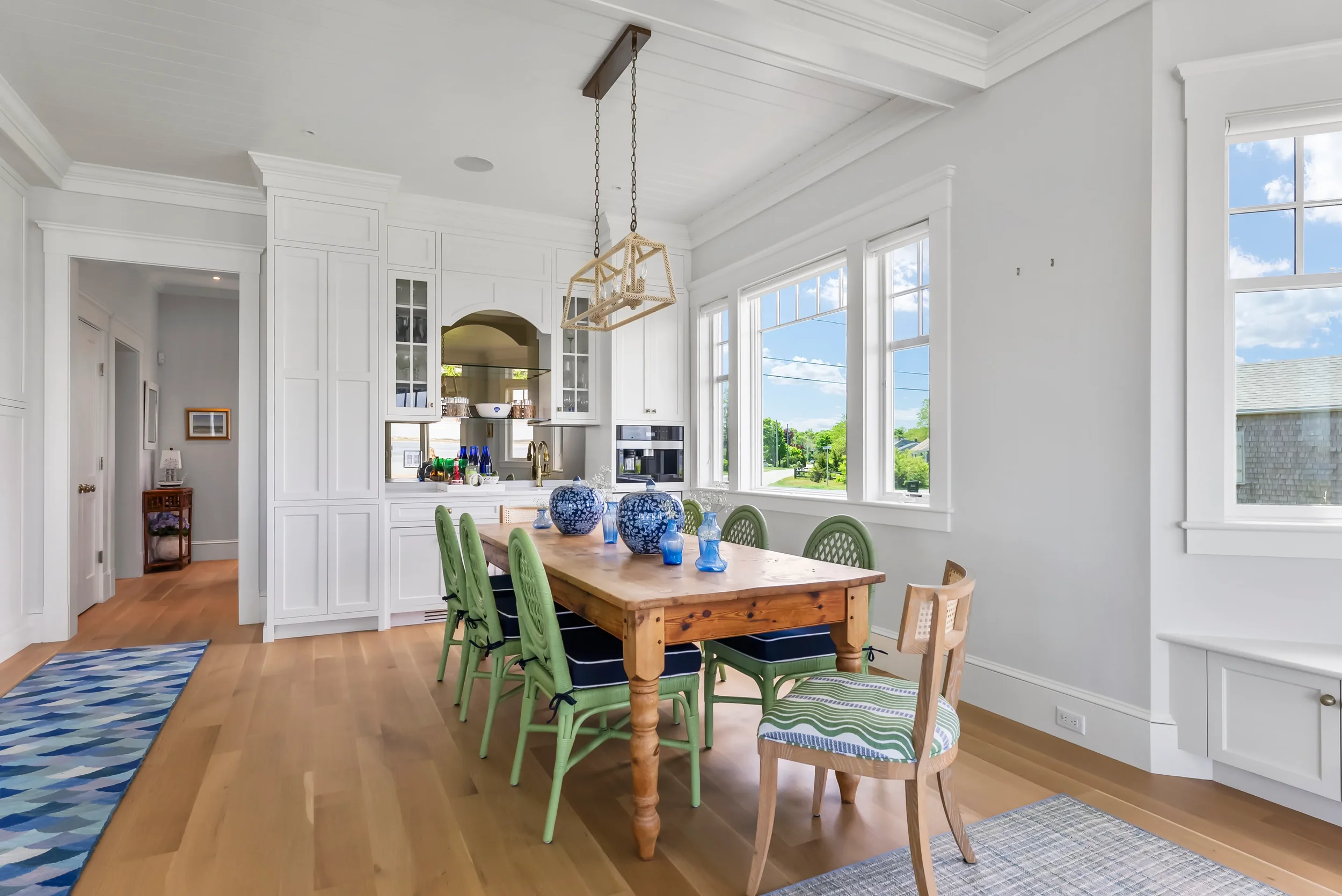 Dining room with a wooden table and eight chairs, four green, one with a striped cushion, and a single wooden chair at the end. Decor includes blue vases and jars. Large windows with a view of a neighborhood street and trees, white walls, hardwood fl