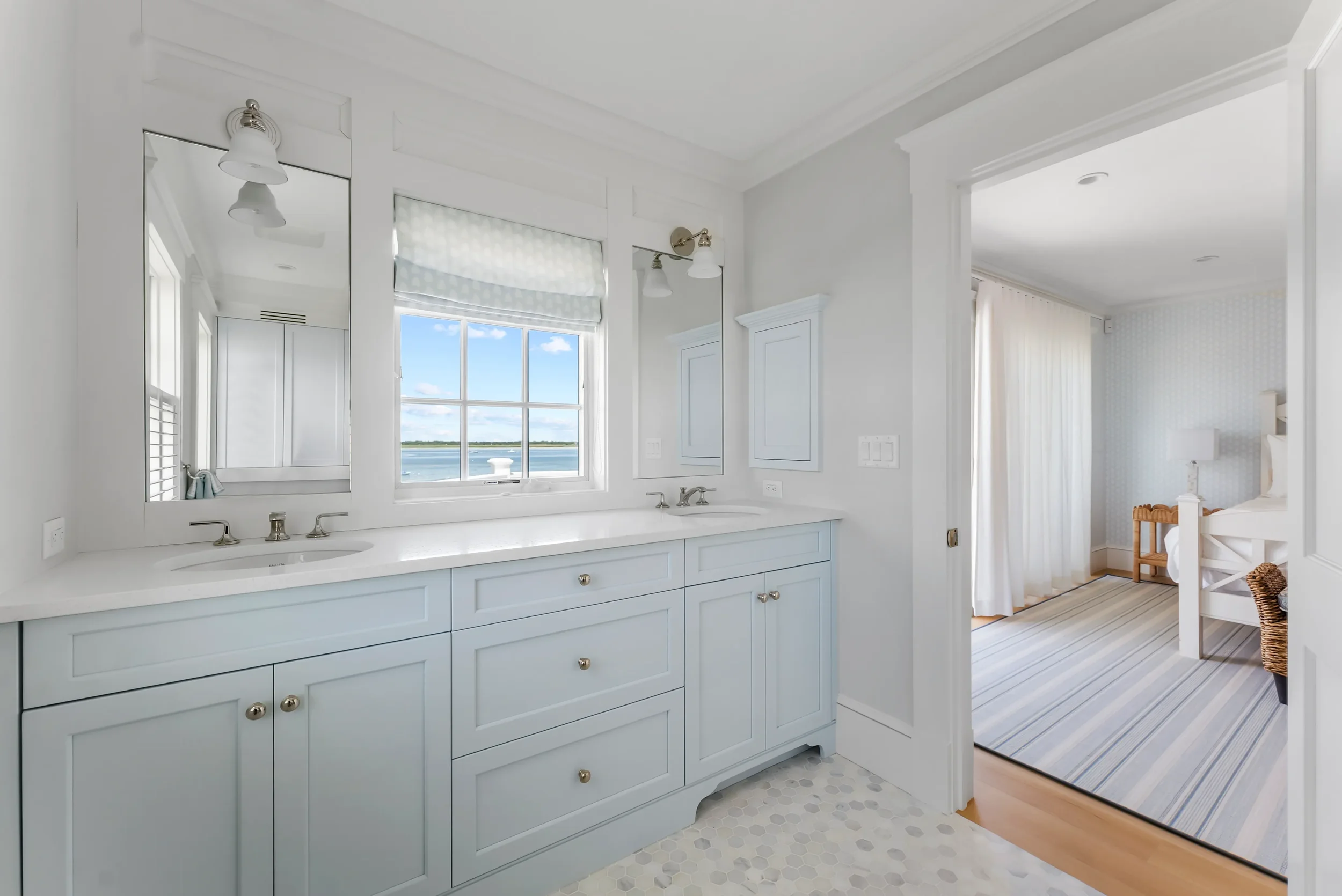 Bathroom with double vanity, large window with view of water, and adjacent bedroom with white bed and striped rug.