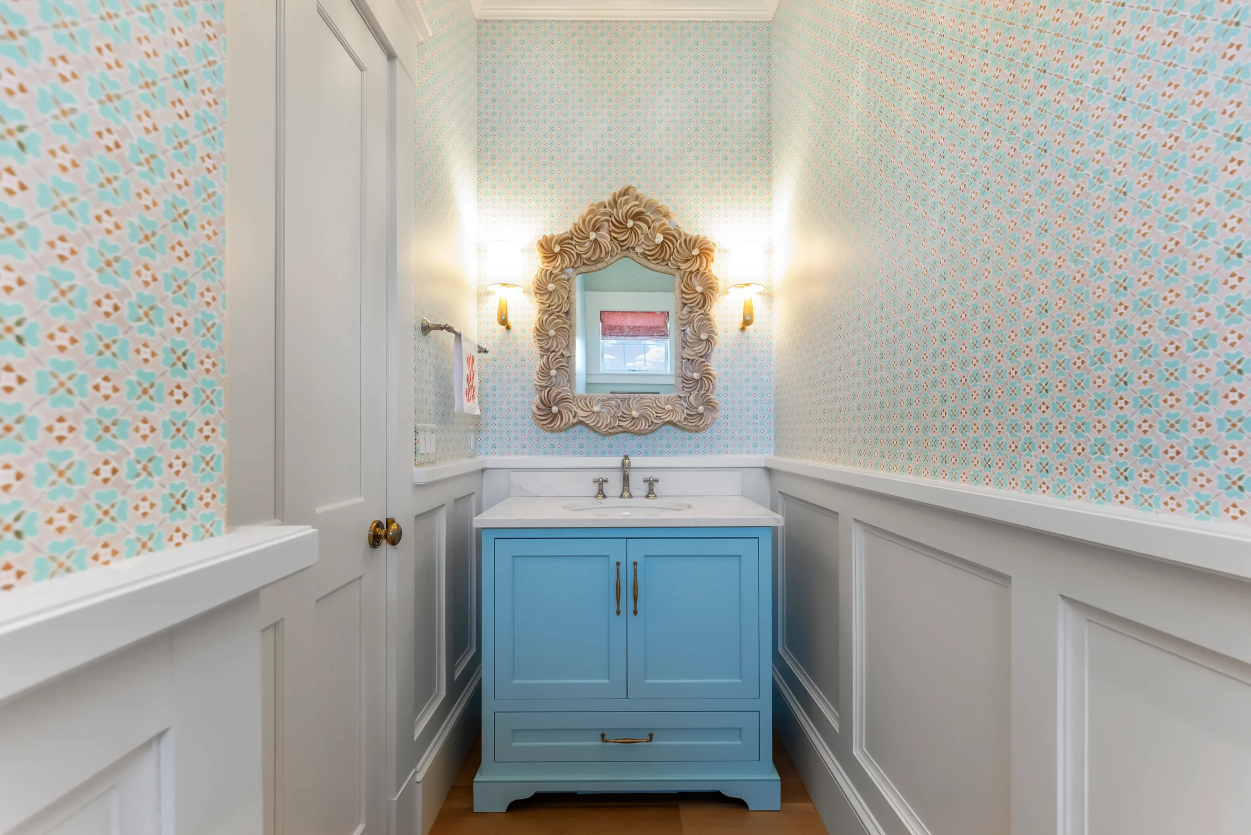 A small bathroom with patterned wallpaper, a light blue vanity with a white countertop and brass handles, a decorative mirror with a shell-like frame, and two wall-mounted sconces on either side of the mirror.